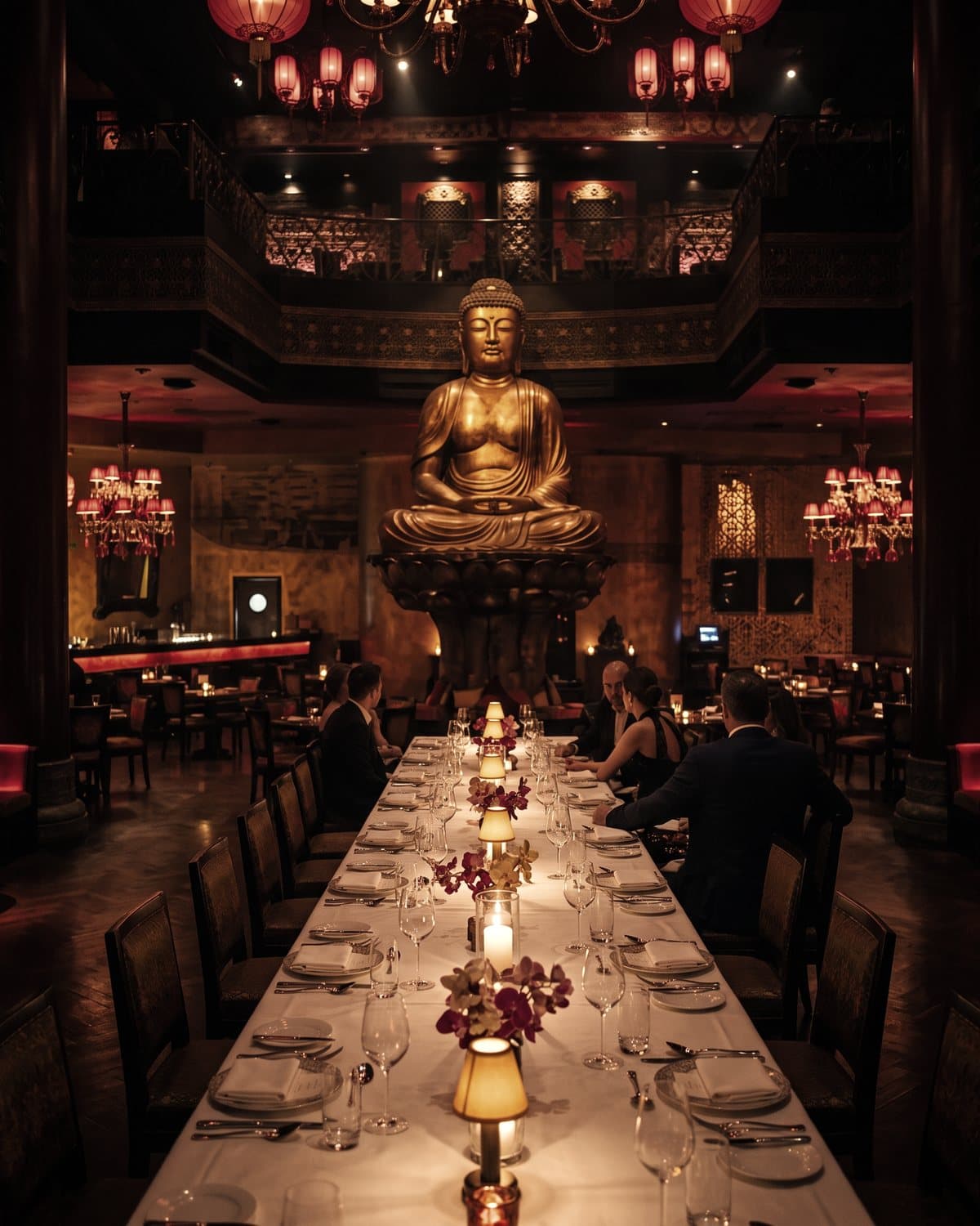 Symbolbild: eine lange, festlich gedeckte Tafel mit Kerzen vor einer großen goldenen Buddha-Statue in einem rot beleuchteten Restaurant