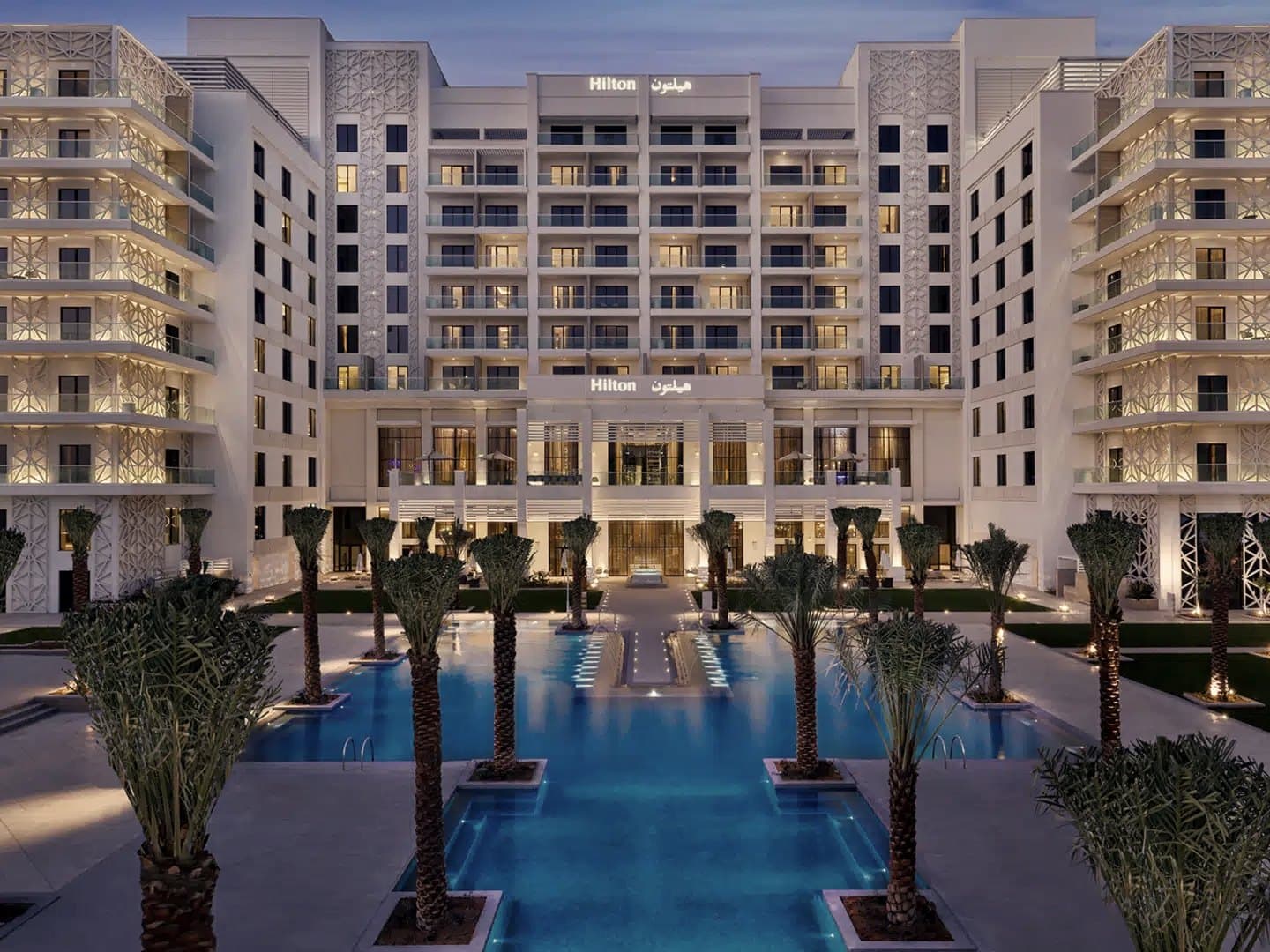 Das Hilton Abu Dhabi Yas Island am Abend: beleuchtete Fassade, Palmen und Pool